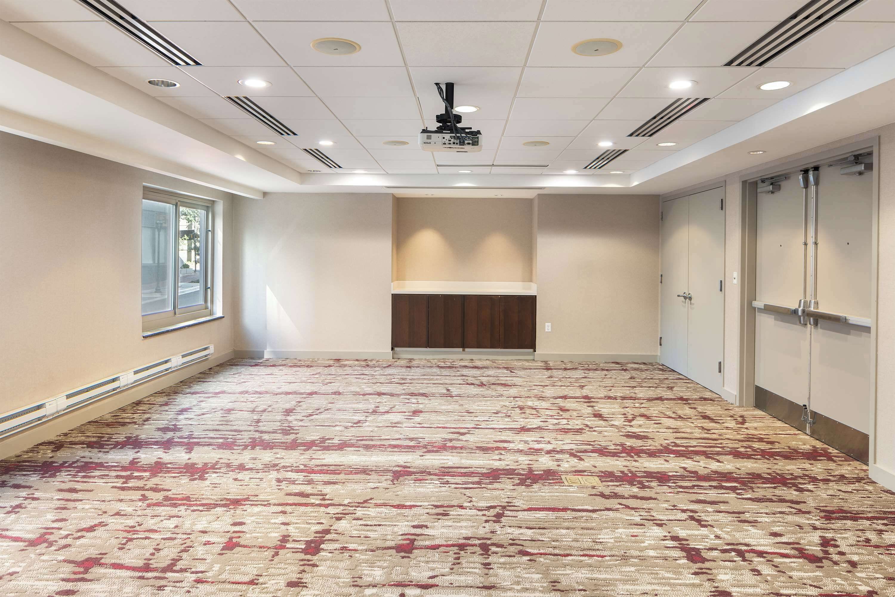 A spacious hotel room with a patterned carpet, a projector ceiling, and a large window on the left.
