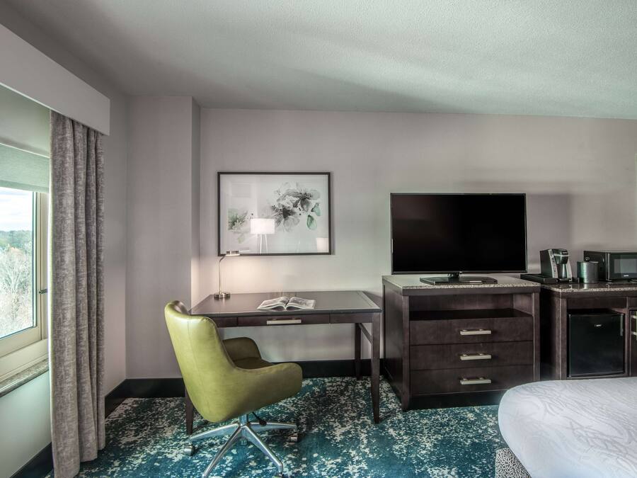 A hotel room with a desk, chair, TV, minibar, and a bed.