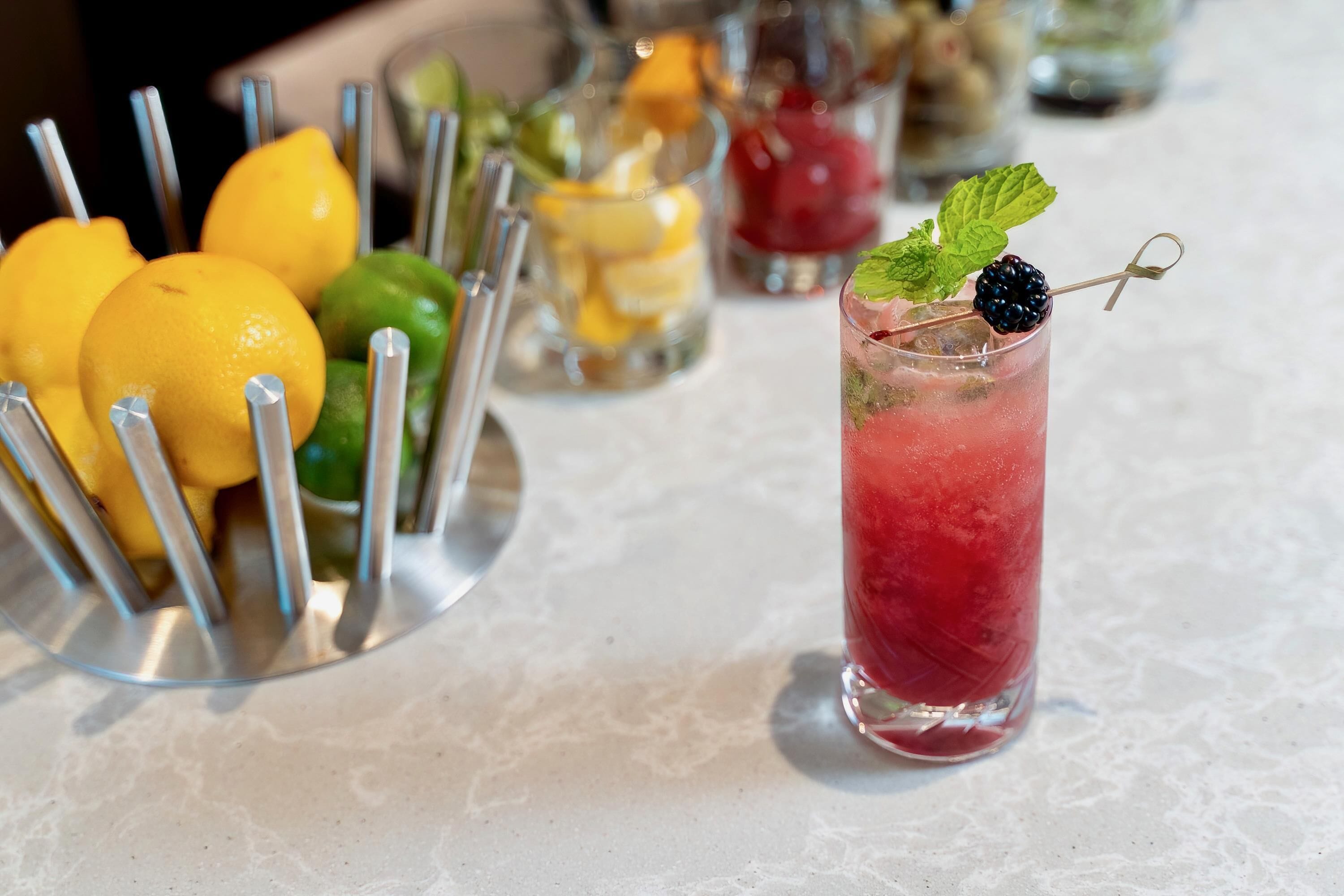 Una zona de bar con diversas bebidas, incluyendo un vaso alto con una bebida roja y una decoración de mora azul, y una bandeja de metal con limones y limas.