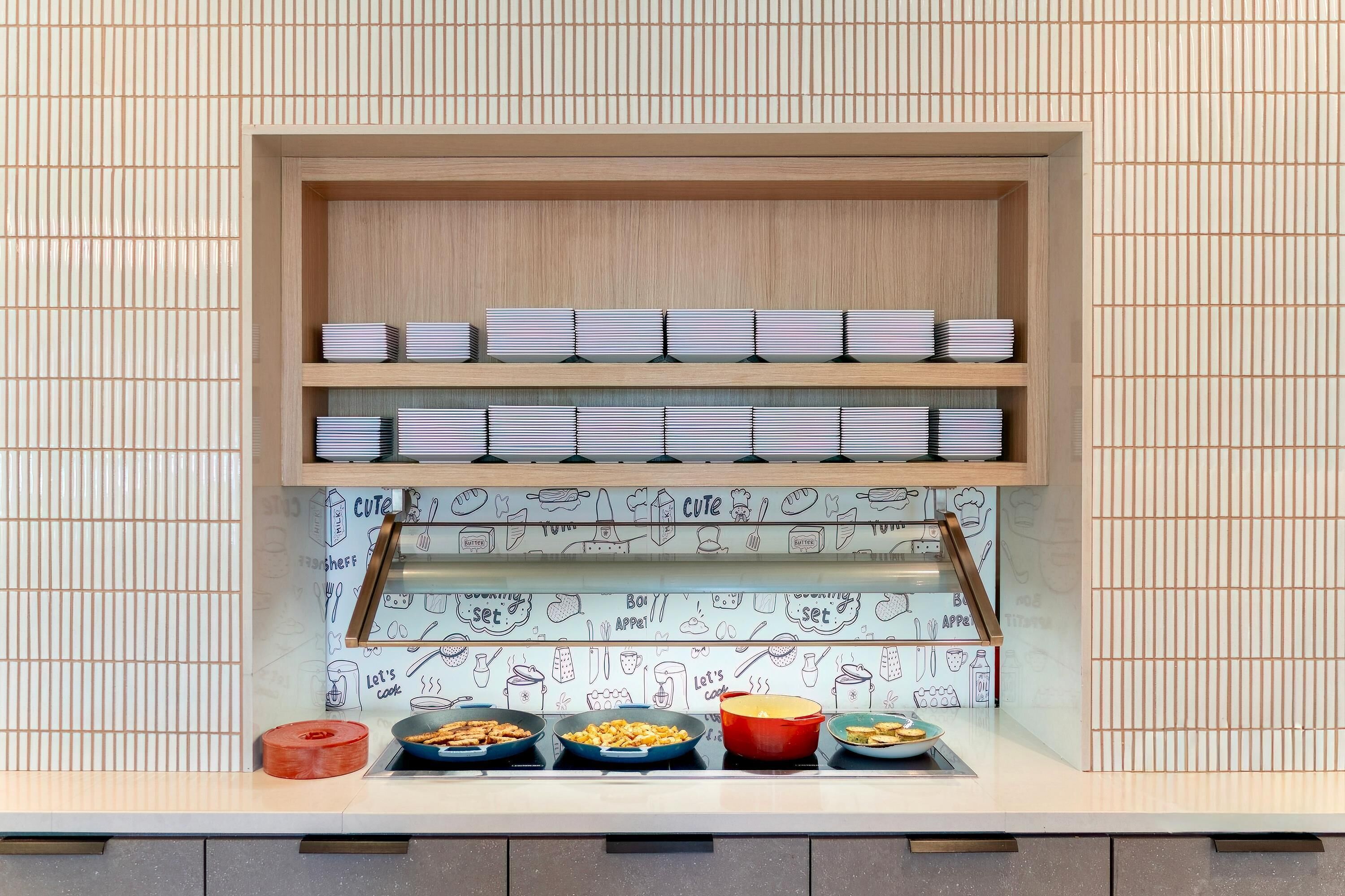 A kitchen counter with a wooden shelf above it, holding neatly stacked plates. Below the shelf, there are three bowls with food, and a red pot on the counter.