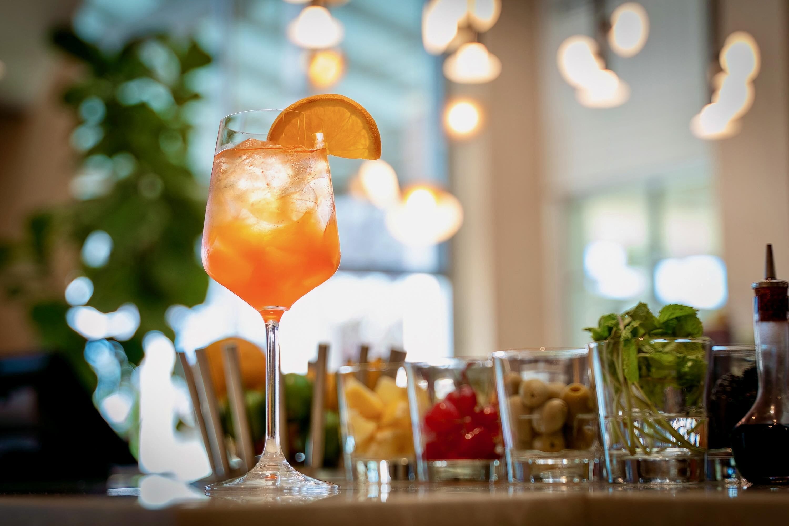 A bar setting with a glass of iced drink garnished with an orange slice, surrounded by various small glasses containing different beverages and garnishes.