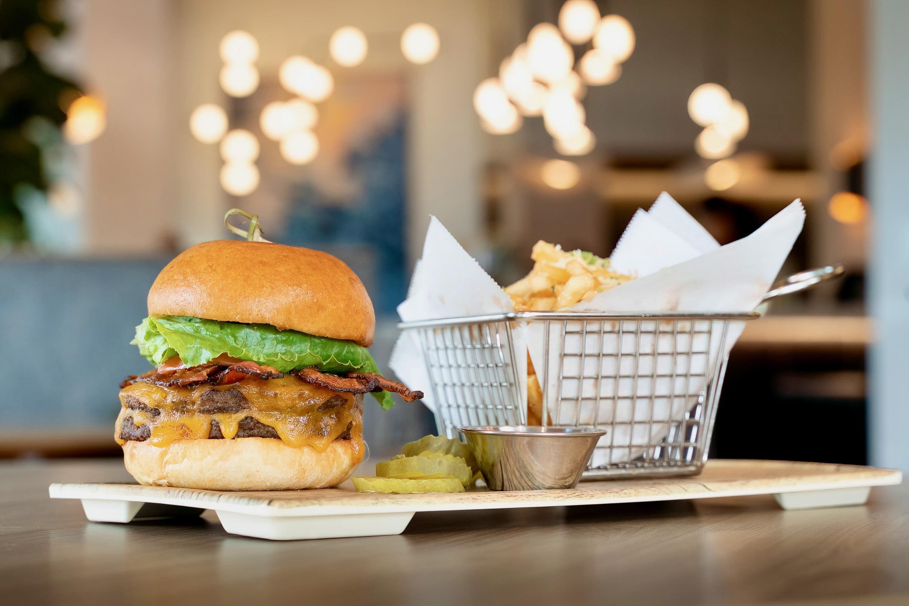 Una hamburguesa con tocino, queso y lechuga en un pan, acompañada de papas fritas y pepinillos sobre una mesa de madera.