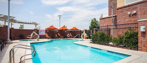 A swimming pool with a sunshade, lounge chairs, and a brick building in the background.