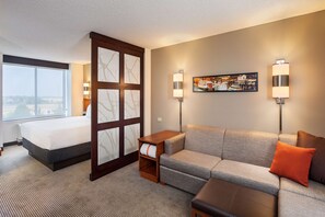 A hotel room with a bed, a sofa, a small table, a wall-mounted TV, and two wall sconces. - Hyatt Place Sacramento Roseville (Roseville)