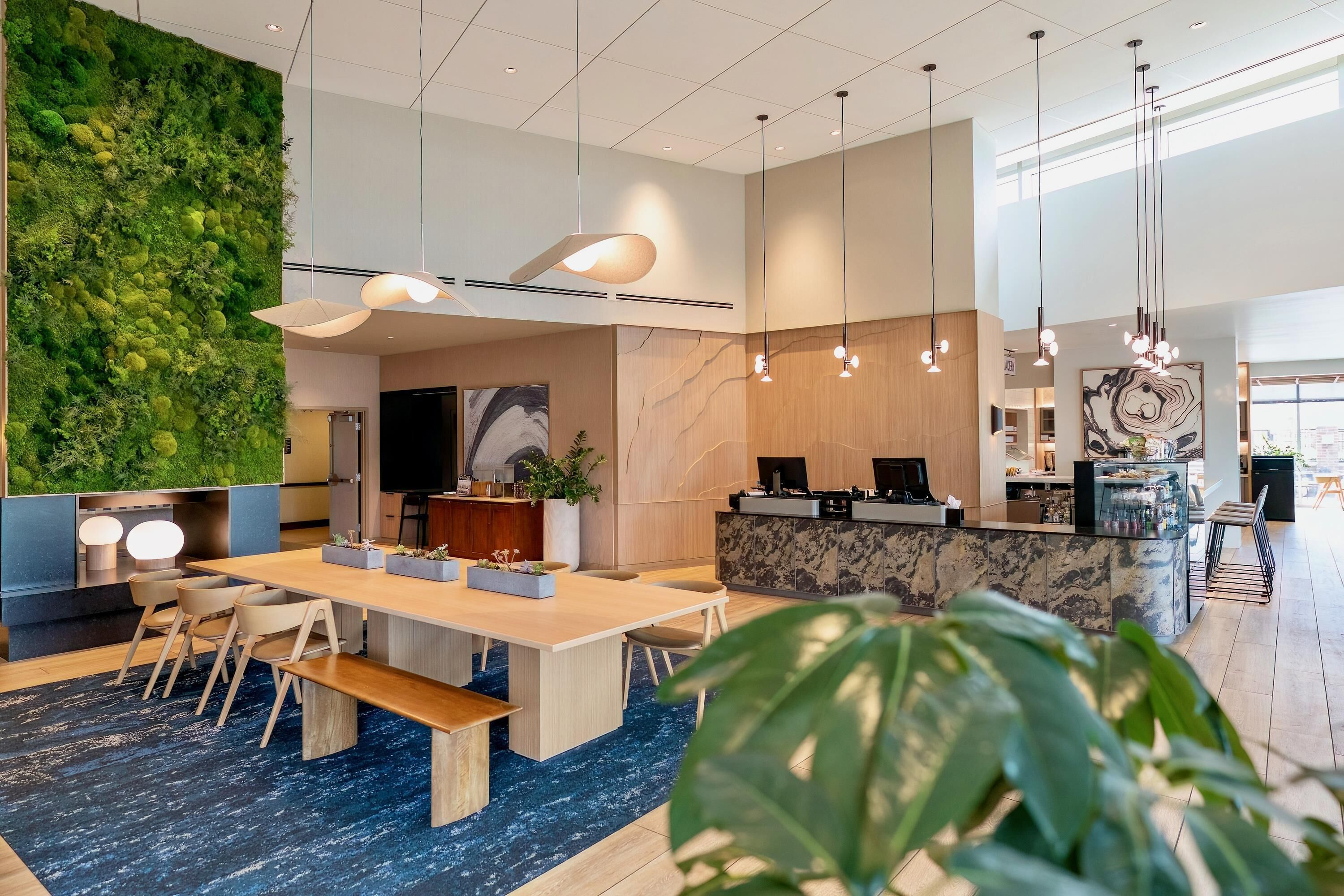 A modern hotel lobby with a green wall, a reception desk, a large TV, and a dining area with a blue carpet.