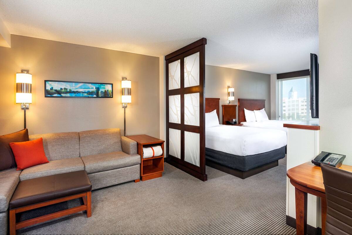 A hotel room with a sofa, a small table, a bedside table, a bed, a nightstand, and a window with a view of buildings.
