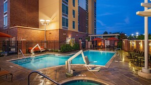 Un espace piscine d’hôtel avec un bâtiment en briques, des chaises longues et un espace salon abrité.