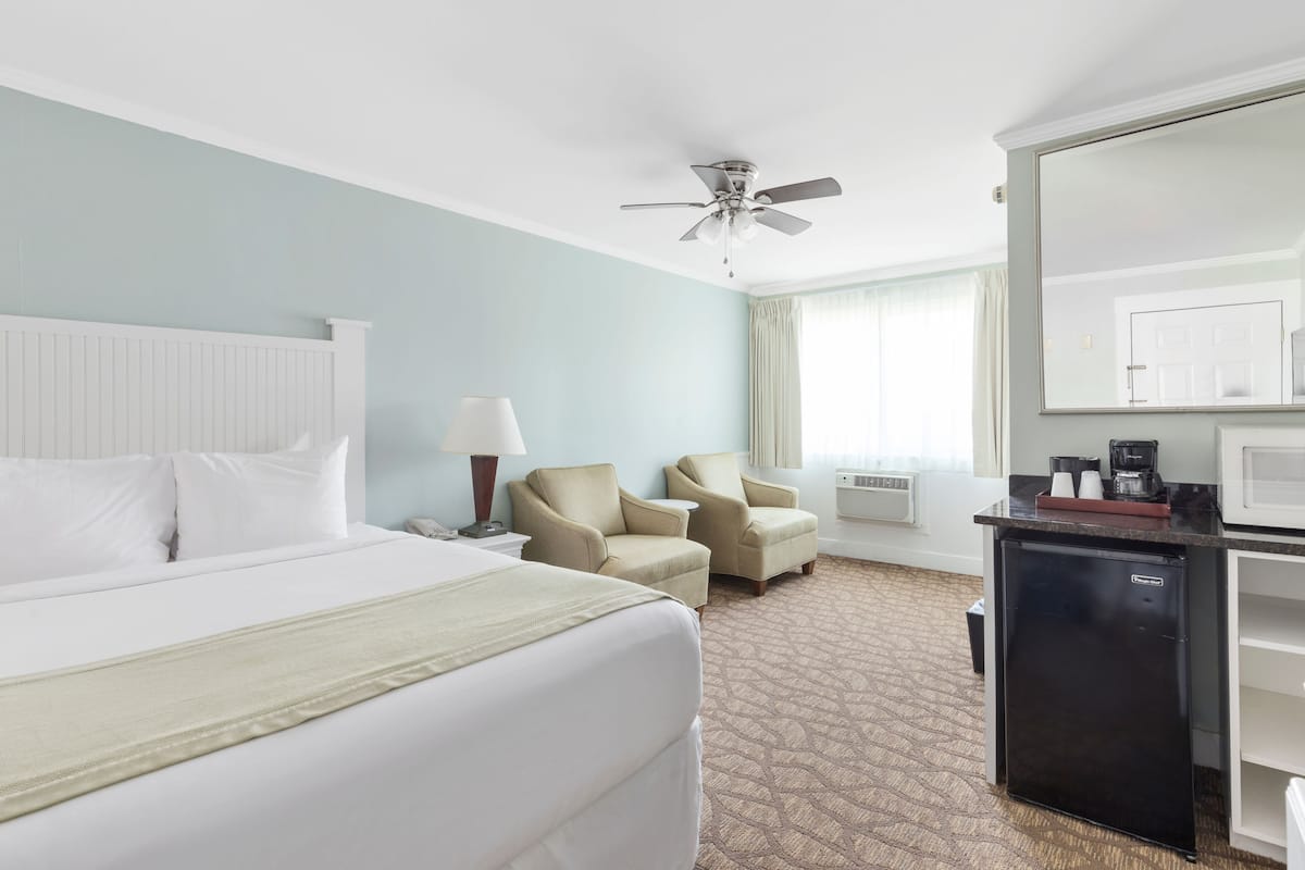 A hotel room with a bed, two armchairs, a ceiling fan, a small refrigerator, a microwave, and a window with curtains.