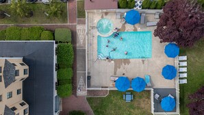 Vista aérea de un área de piscina con sillones de descanso y sombrillas, rodeada de vegetación y un edificio.