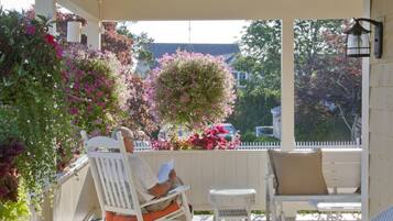 A covered porch with rocking chairs and a table, surrounded by potted plants and flowers.
