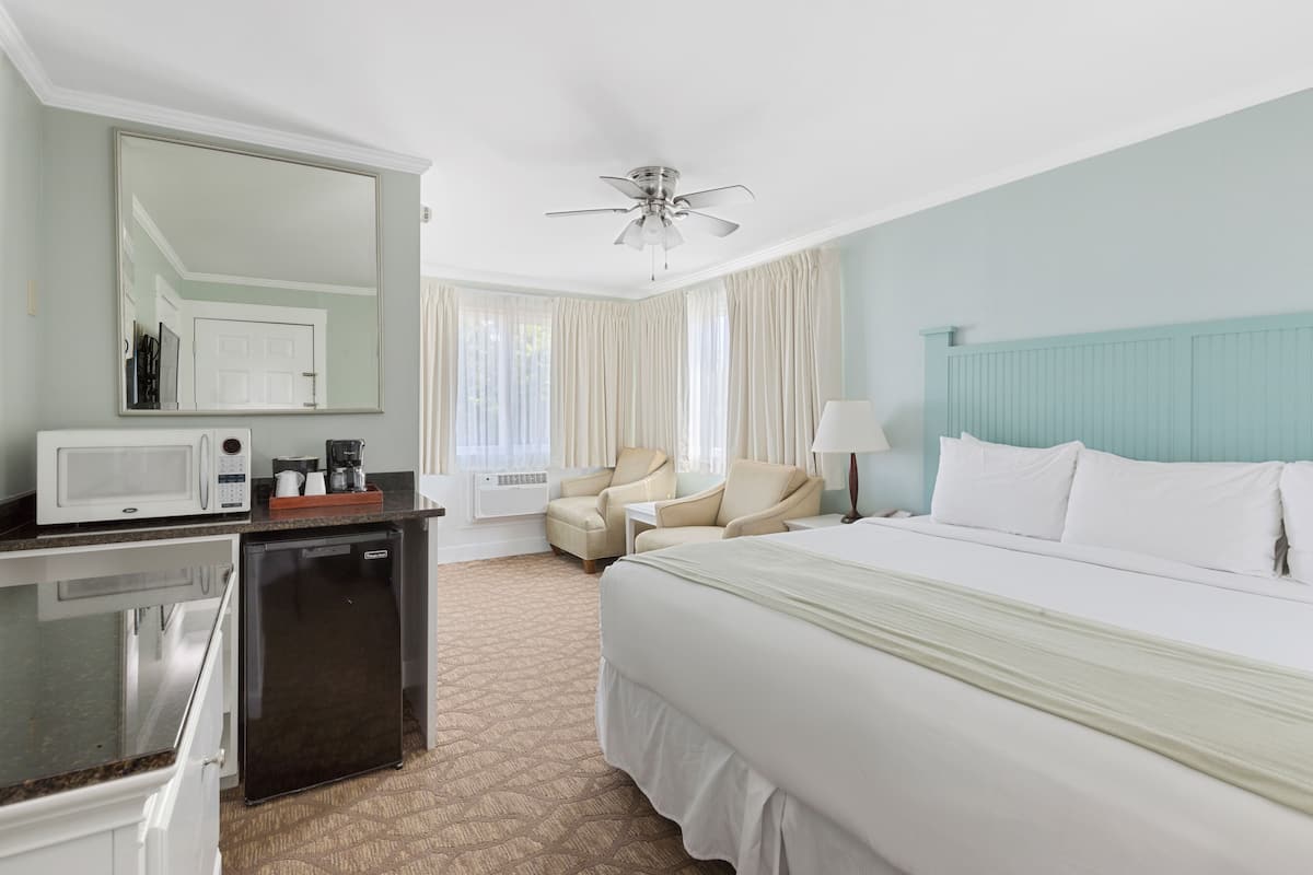 A hotel room with a bed, a desk with a microwave, a refrigerator, a ceiling fan, and a large window with curtains.