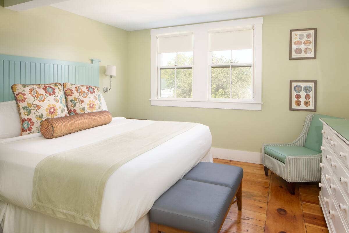 A bedroom with a bed, bench, dresser, chair, and a window with blinds.