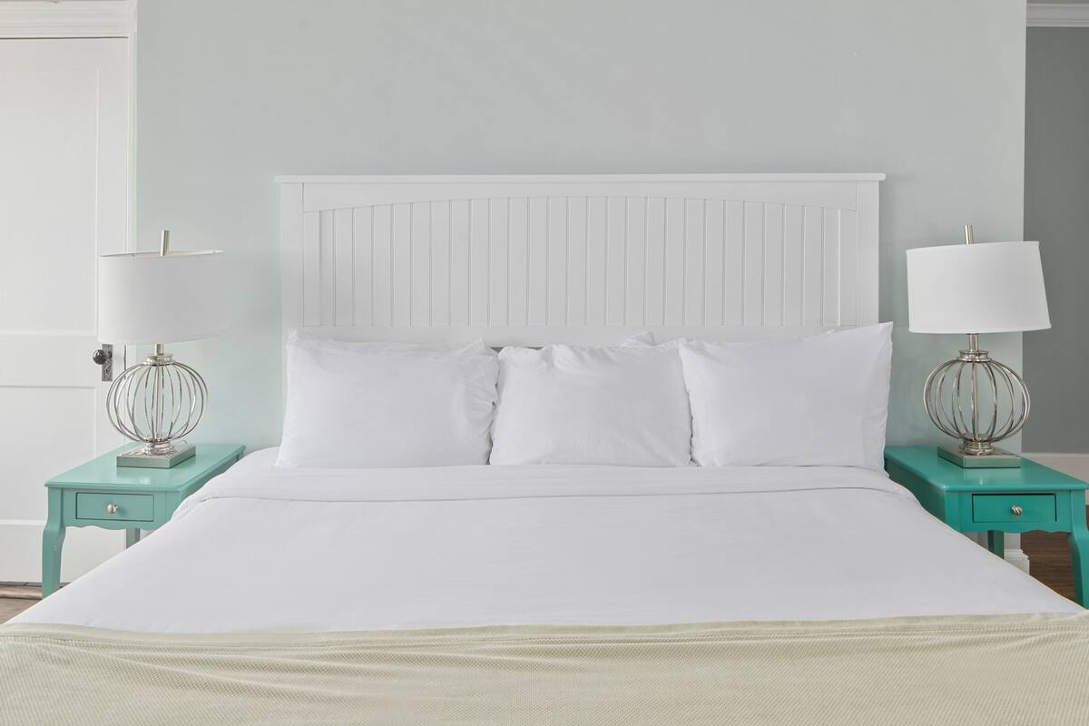 A neatly made bed with white linens, flanked by two teal nightstands with lamps and a drawer, against a light blue wall.