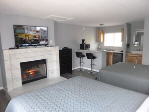 A modern hotel room with a fireplace, a television mounted above it, a kitchen area with a microwave and coffee maker, and a seating area with two bar stools.