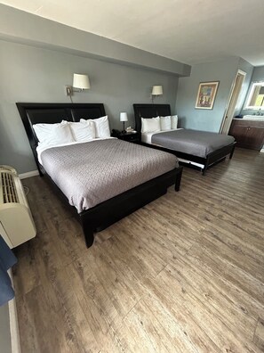 A hotel room with two beds, a wooden floor, a bathroom with a sink, and a painting on the wall.