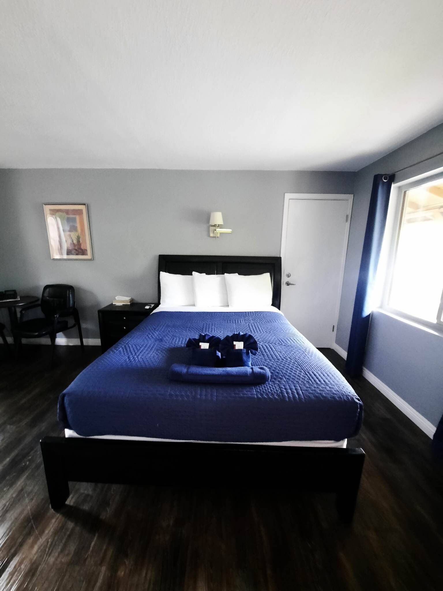Un lit bien fait, recouvert d’une couverture bleue, une lampe de chevet, un tableau encadré accroché au mur et un bureau avec une chaise.