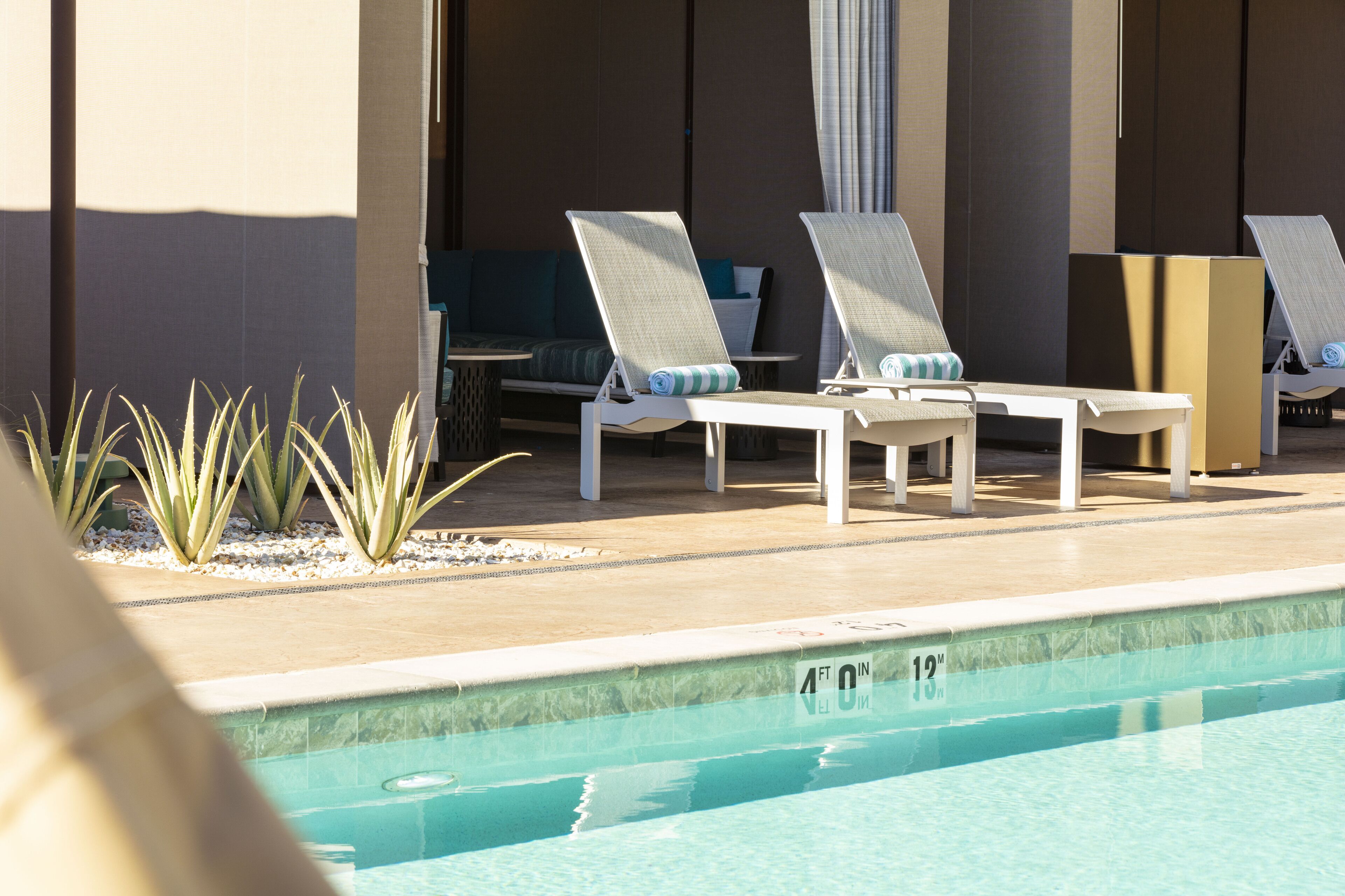 Zona de piscina con sillones de descanso y una mesita, piscina con indicador de profundidad, y una planta en primer plano.