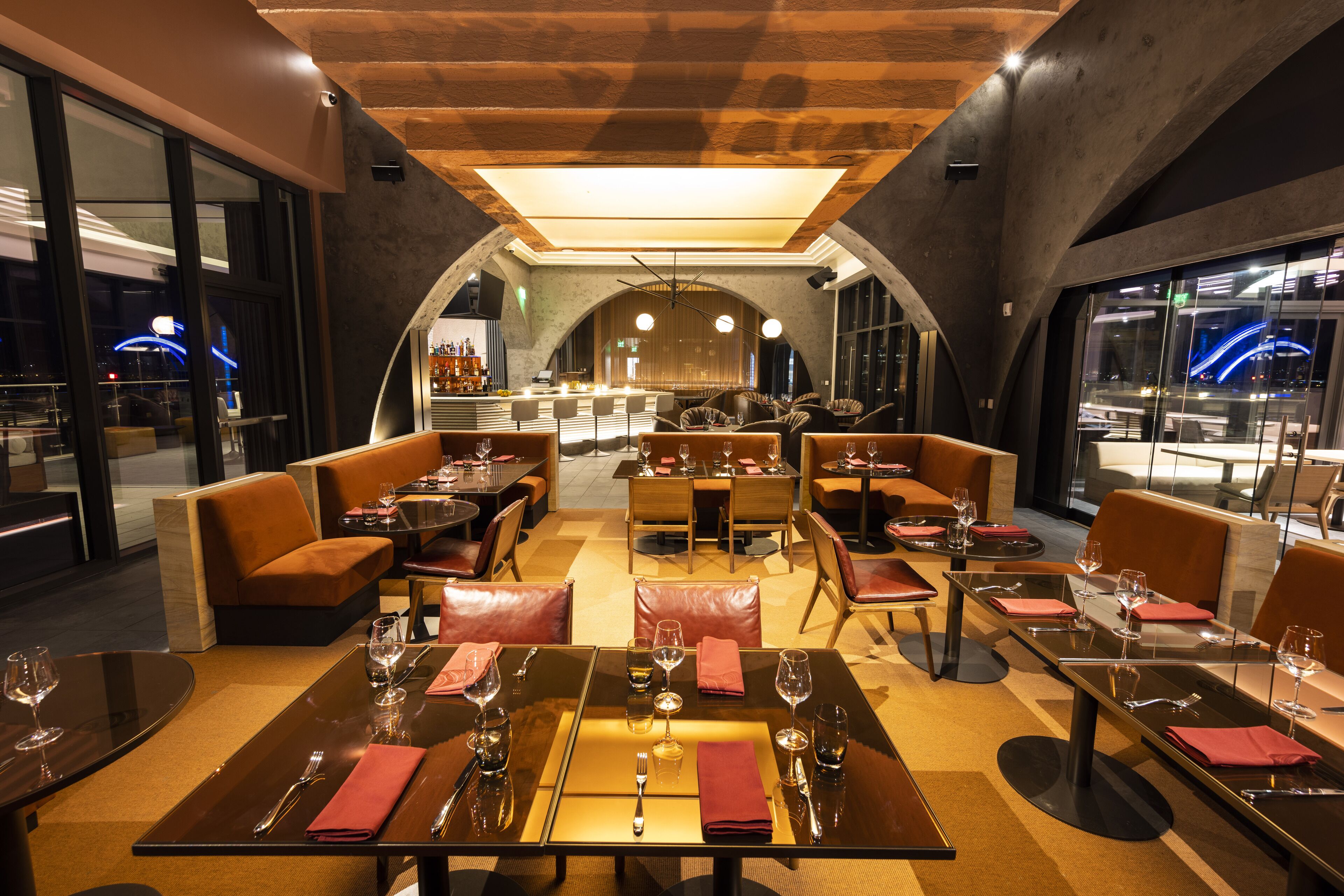 Un restaurante moderno con mesas altas y cabinas, una zona de bar y grandes ventanales con vistas a la ciudad.