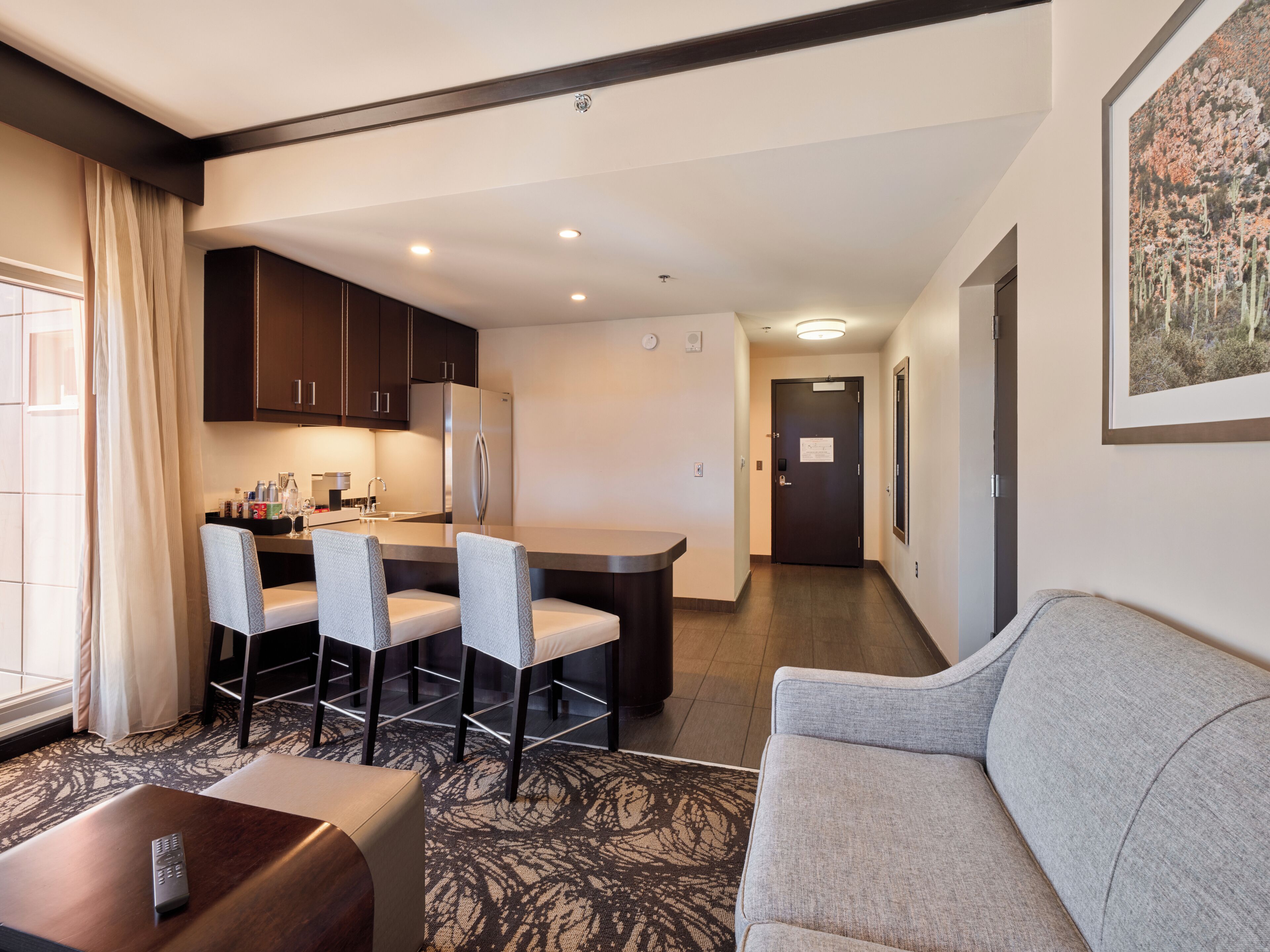 Una habitación de hotel moderna con kitchenette, zona de comedor, sofá y un control remoto sobre una mesa.