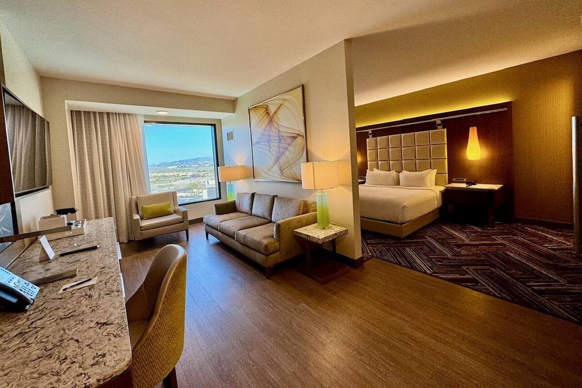 Habitación de hotel con cama, escritorio, silla, sofá, lámpara y vistas al exterior.