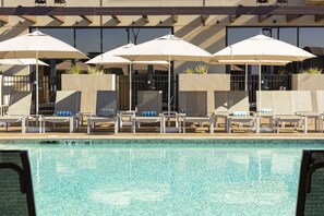 Zona de piscina con sillas de descanso y sombrillas, una piscina cristalina y un edificio con grandes ventanales al fondo.