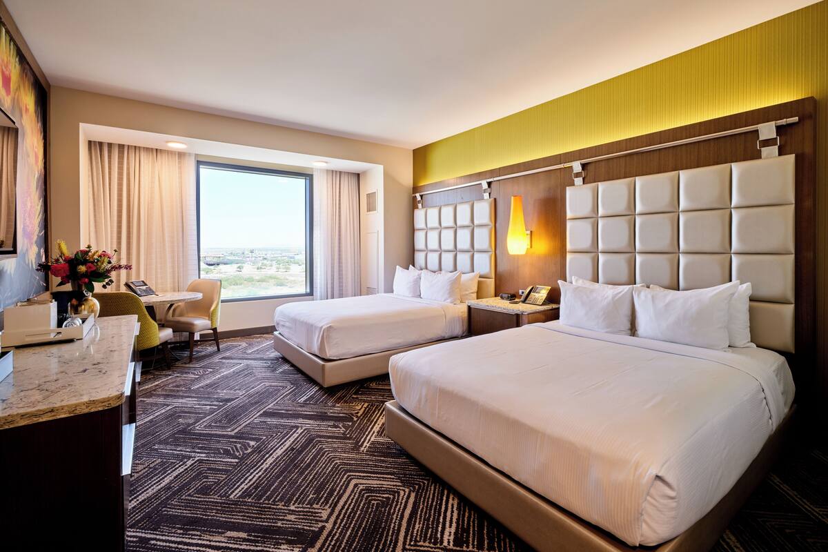 Habitación de hotel con dos camas, un escritorio, una silla, un ventanal con cortinas y vistas a la ciudad.