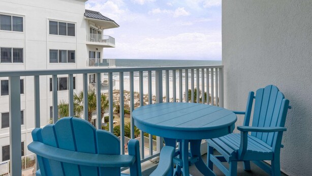 A balcony with a table and two chairs, overlooking a beach and a white building. - Palm Beach Resort Orange Beach a Ramada by Wyndham (Orange Beach)
