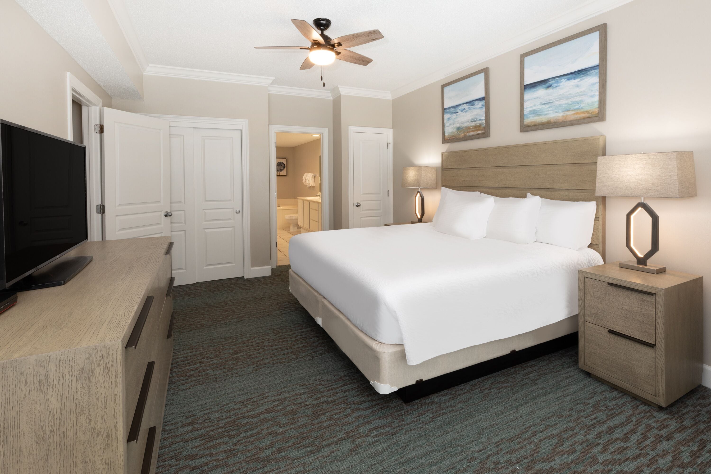 A hotel room with a bed, a TV, a ceiling fan, and artwork on the wall.