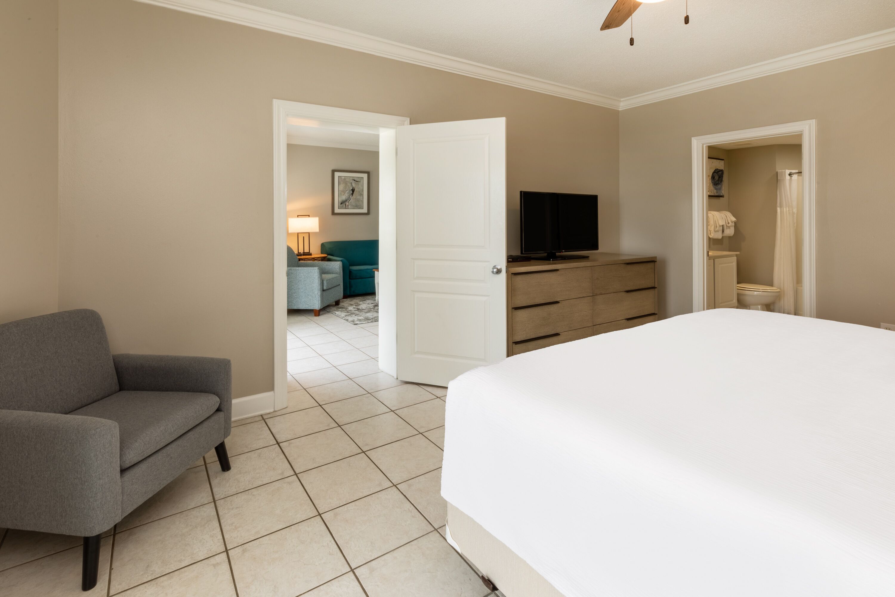 Un dormitorio con cama, un sillón gris, un aparador, televisor y un baño visible a través de una puerta abierta.
