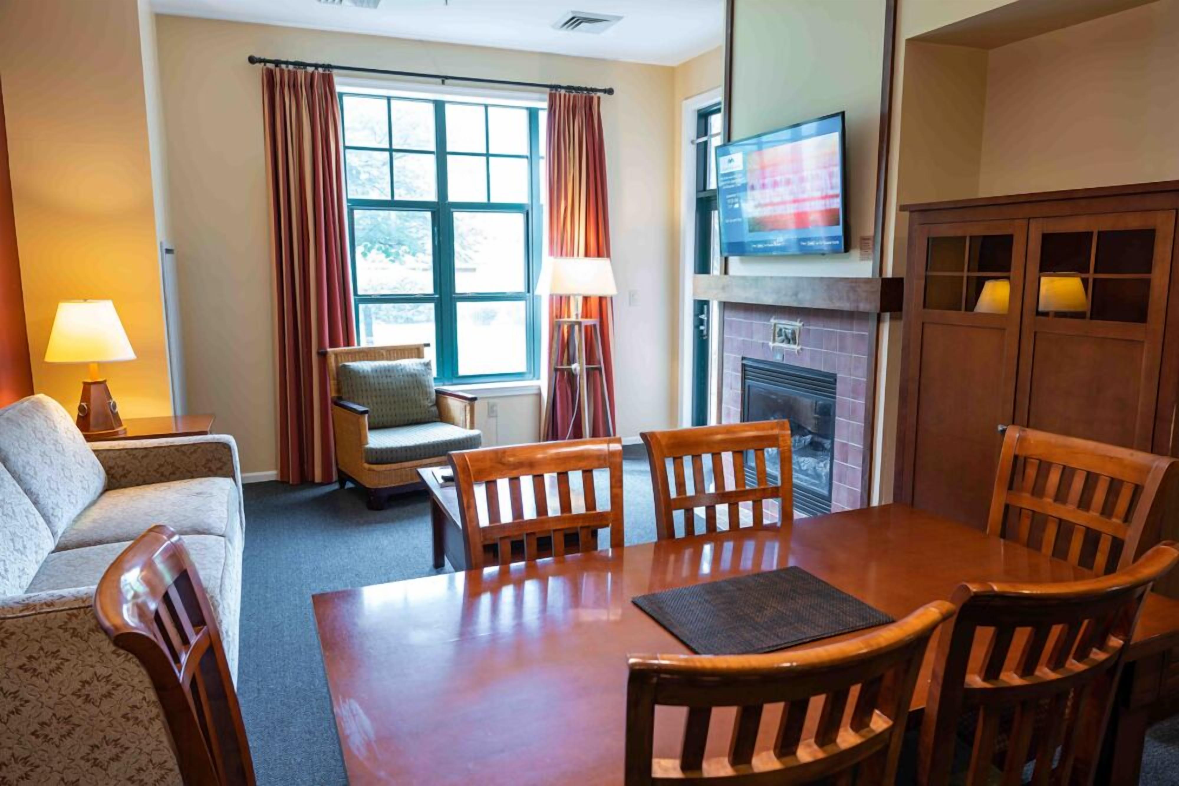 Una habitación de hotel con una mesa de comedor de madera, cuatro sillas, un sofá, un televisor sobre la chimenea y una ventana con cortinas.