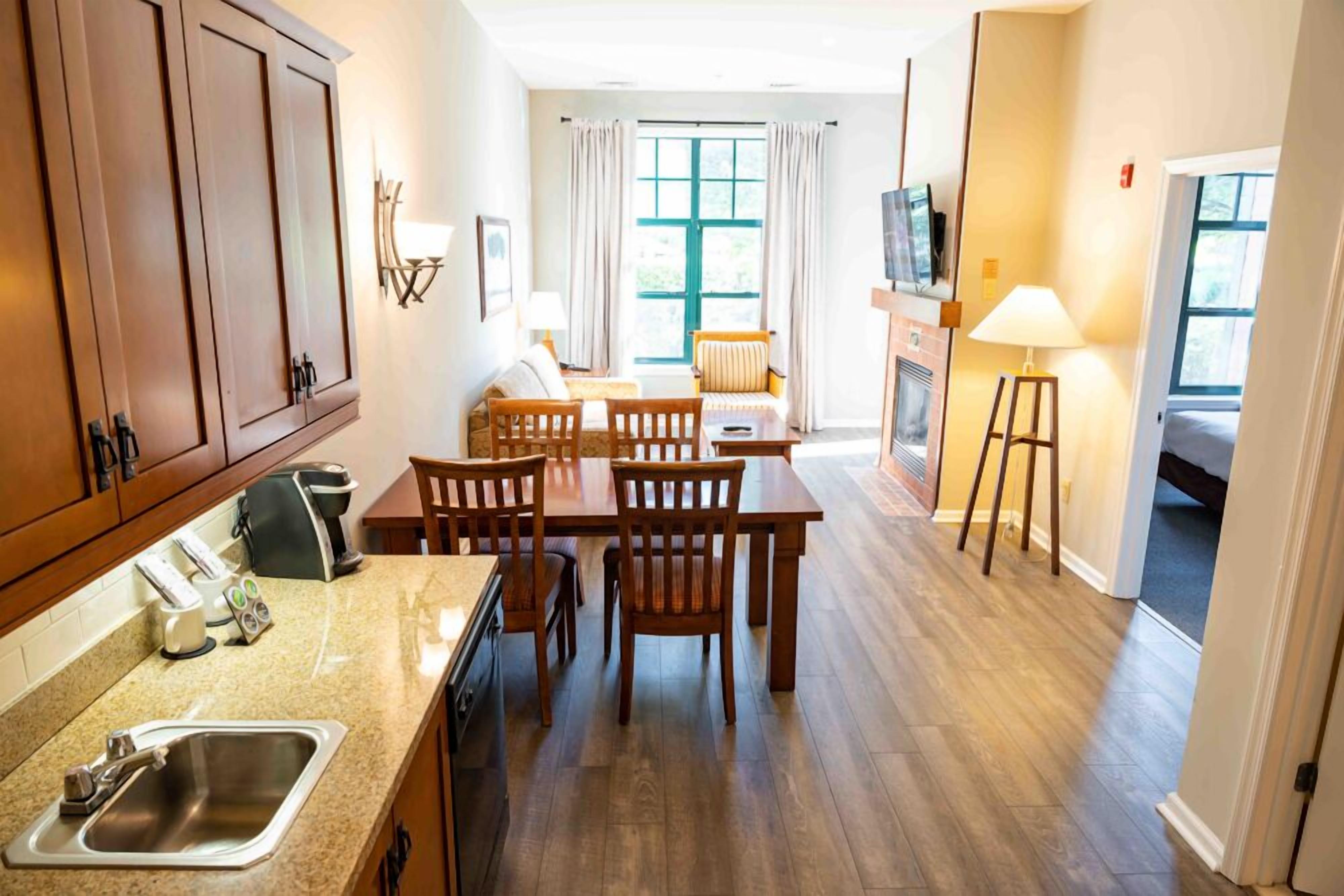 A hotel room with a kitchen area, dining table, chairs, a sofa, a TV, and a lamp.