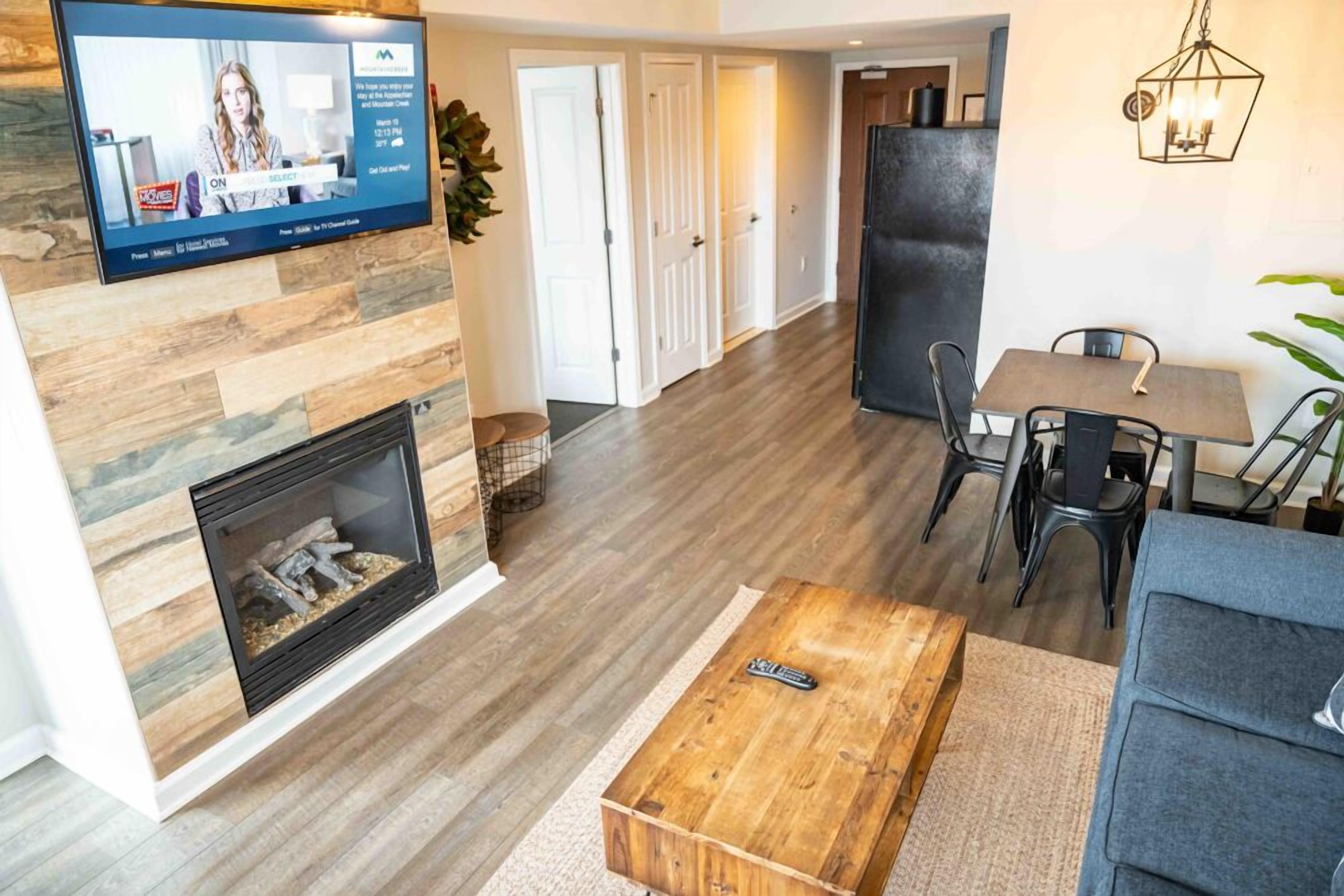 A modern living room with a fireplace, a television mounted on the wall, a dining area with a table and chairs, and a kitchen area in the background.