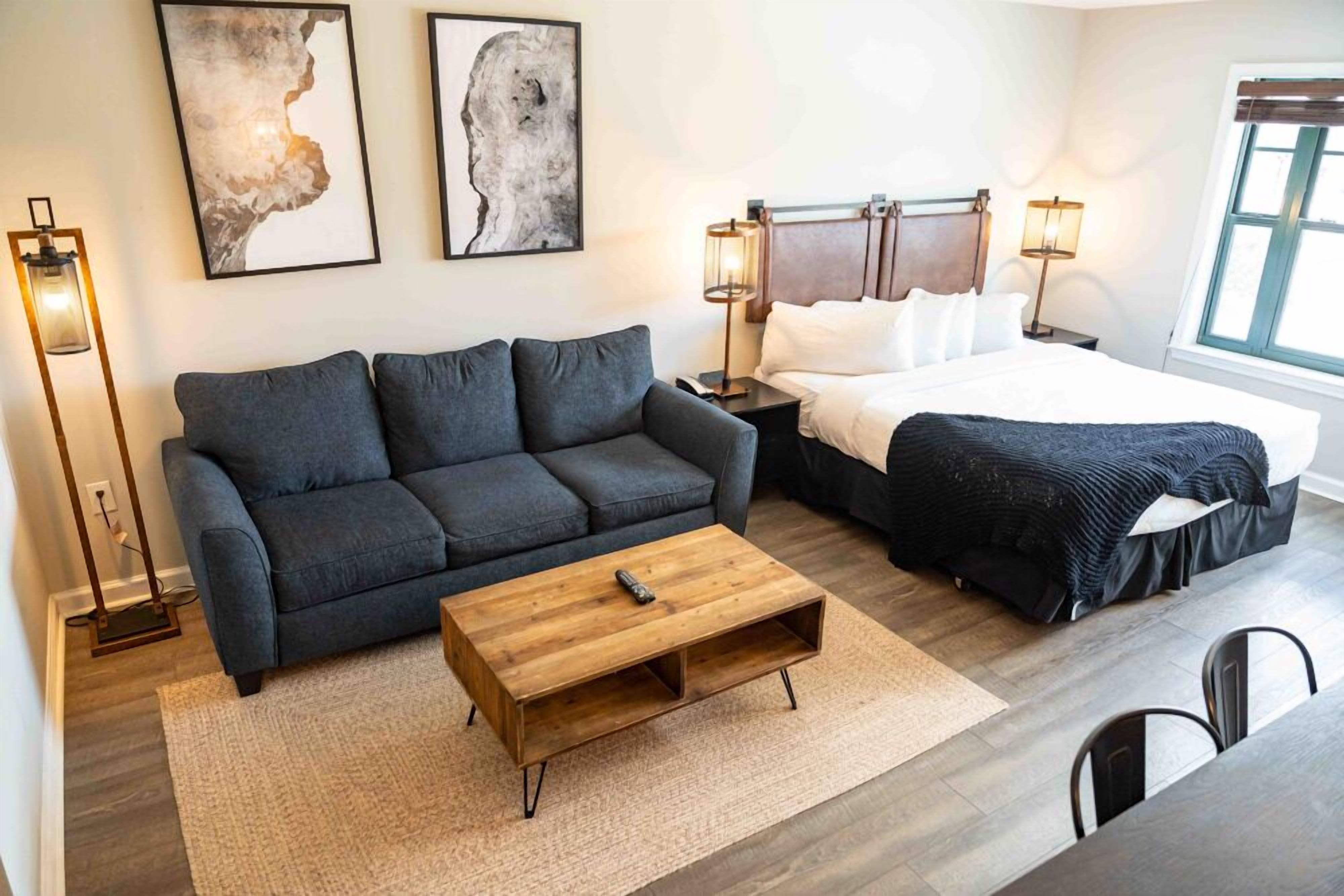 A modern hotel room with a dark gray sofa, a wooden coffee table, a bed with white linens, and two abstract paintings on the wall.