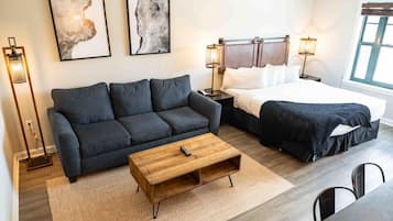 A modern hotel room with a dark gray sofa, a wooden coffee table, a bed with white linens, and two abstract paintings on the wall.