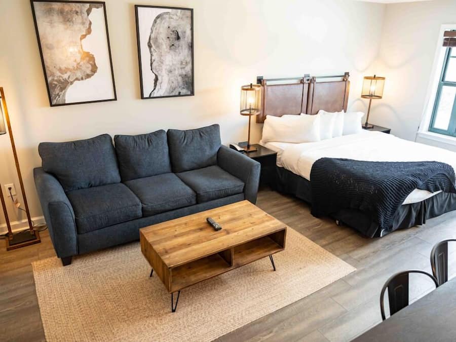 A modern hotel room with a dark gray sofa, a wooden coffee table, a bed with white linens, and two abstract paintings on the wall.
