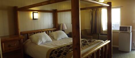 A wooden four-poster bed with a patterned throw blanket, a bedside table, a lamp, and a window with curtains.