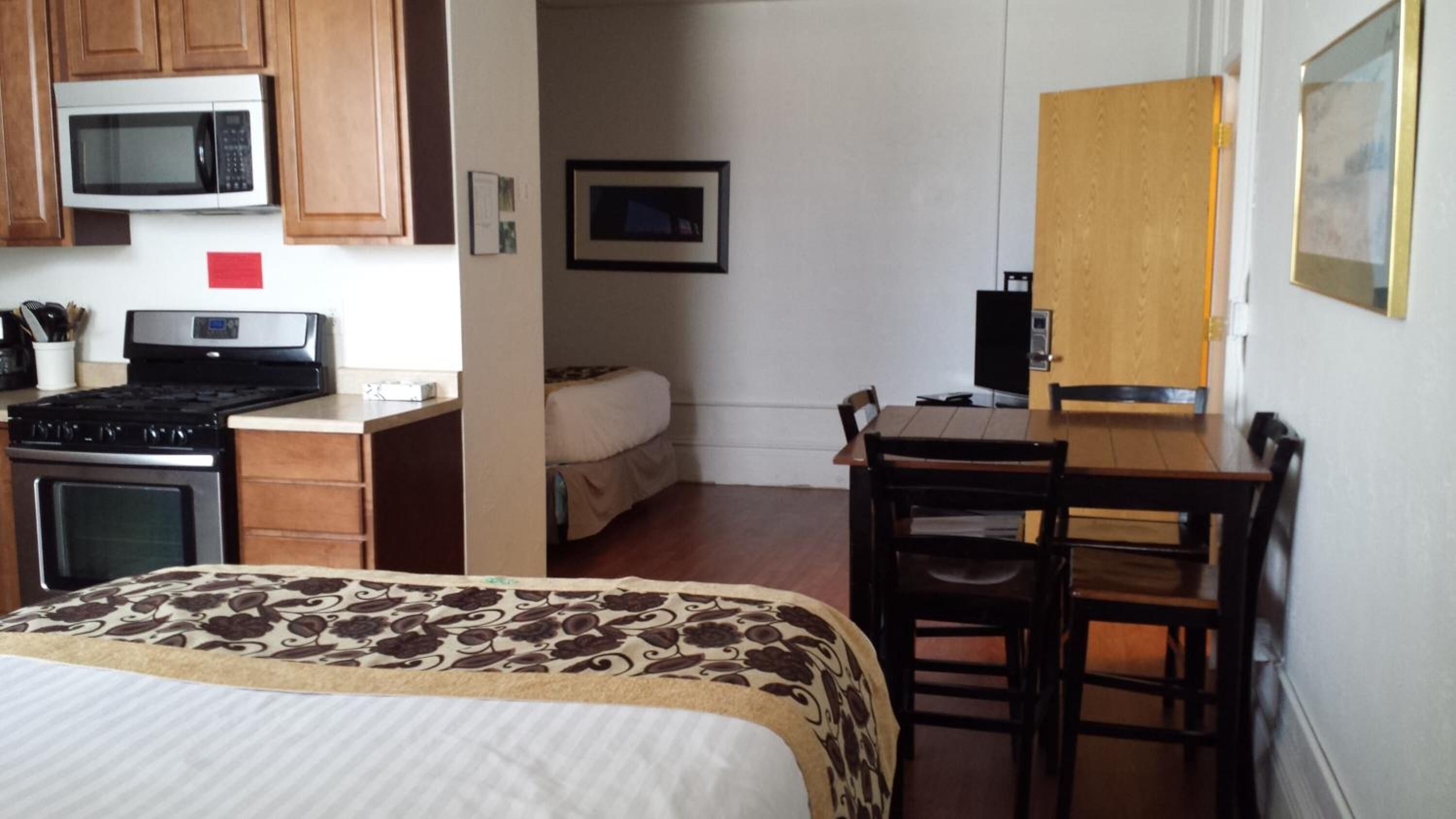 A compact hotel room with a bed, a kitchenette with a stove and microwave, a small dining table with chairs, and a door leading to another room.