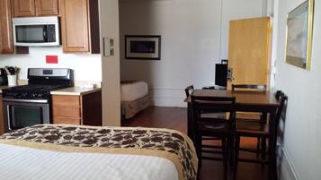 A compact hotel room with a bed, a kitchenette with a stove and microwave, a small dining table with chairs, and a door leading to another room.