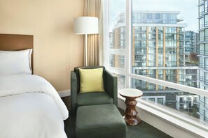 Een hotelkamer met een groot bed, een groene fauteuil met een geel kussen, een klein houten tafeltje en uitzicht op moderne gebouwen door een groot raam.
