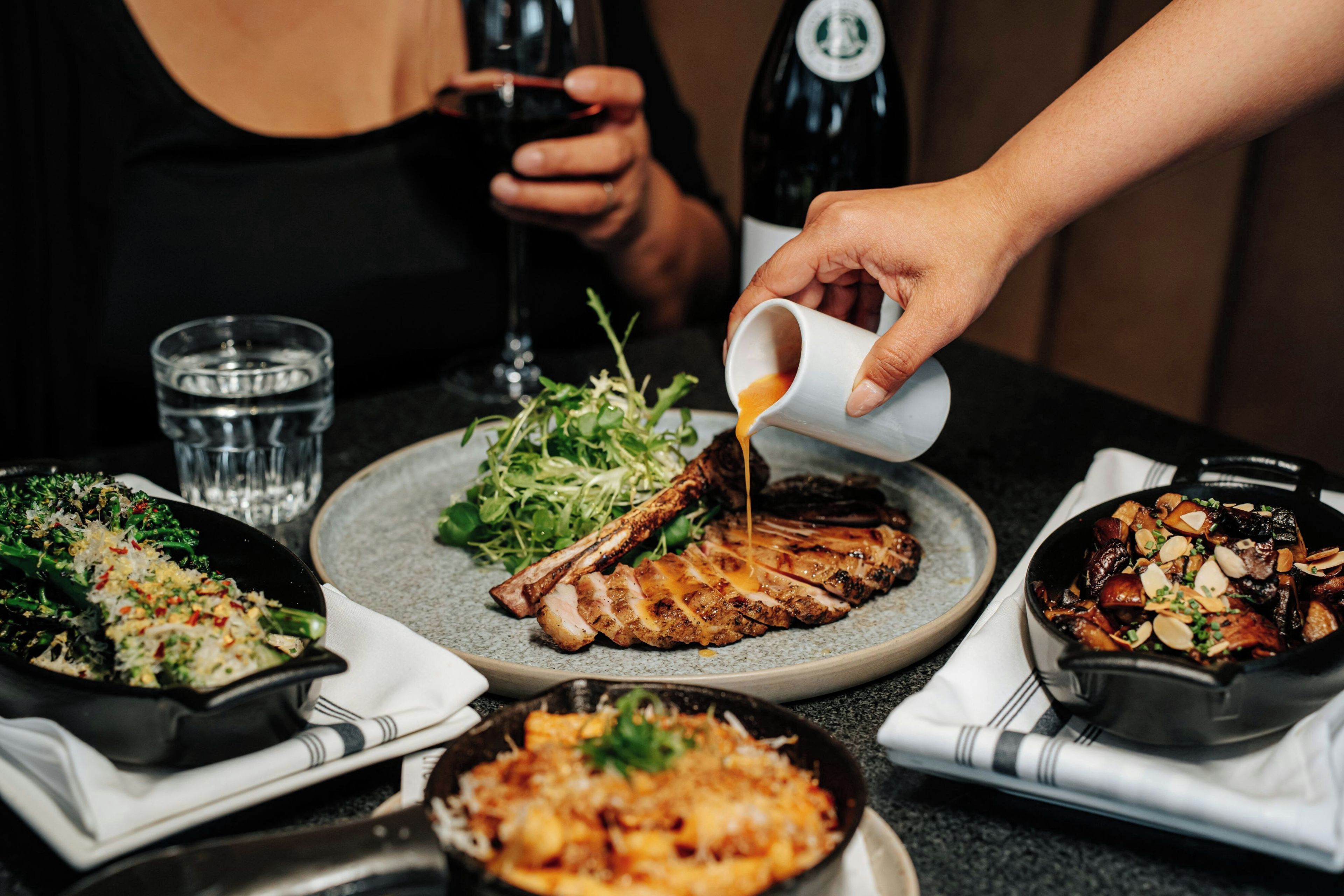 A dining table with various dishes, including a steak, salad, and a bowl of food, with a person pouring sauce over the steak.