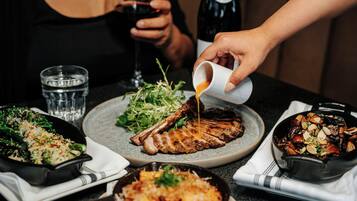 A dining table with various dishes, including a steak, salad, and a bowl of food, with a person pouring sauce over the steak.