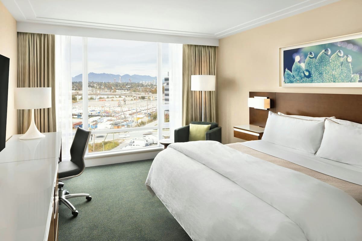 Ein Hotelzimmer mit einem großen Bett, einem Schreibtisch, einem Sessel und Blick auf eine Stadtlandschaft durch große Fenster.