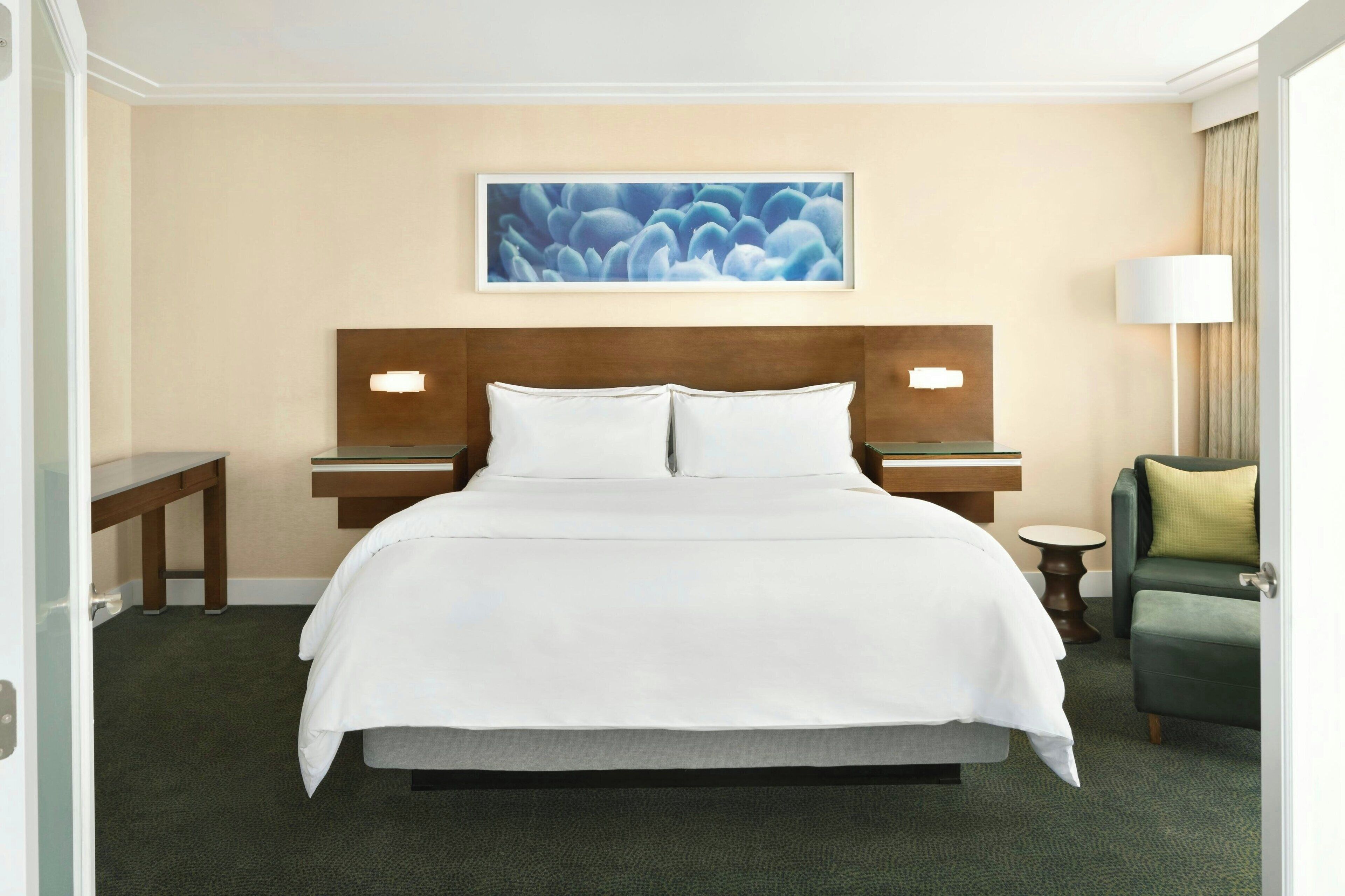Habitación de hotel con una cama grande, dos mesitas de noche, una silla, una lámpara y un cuadro en la pared.