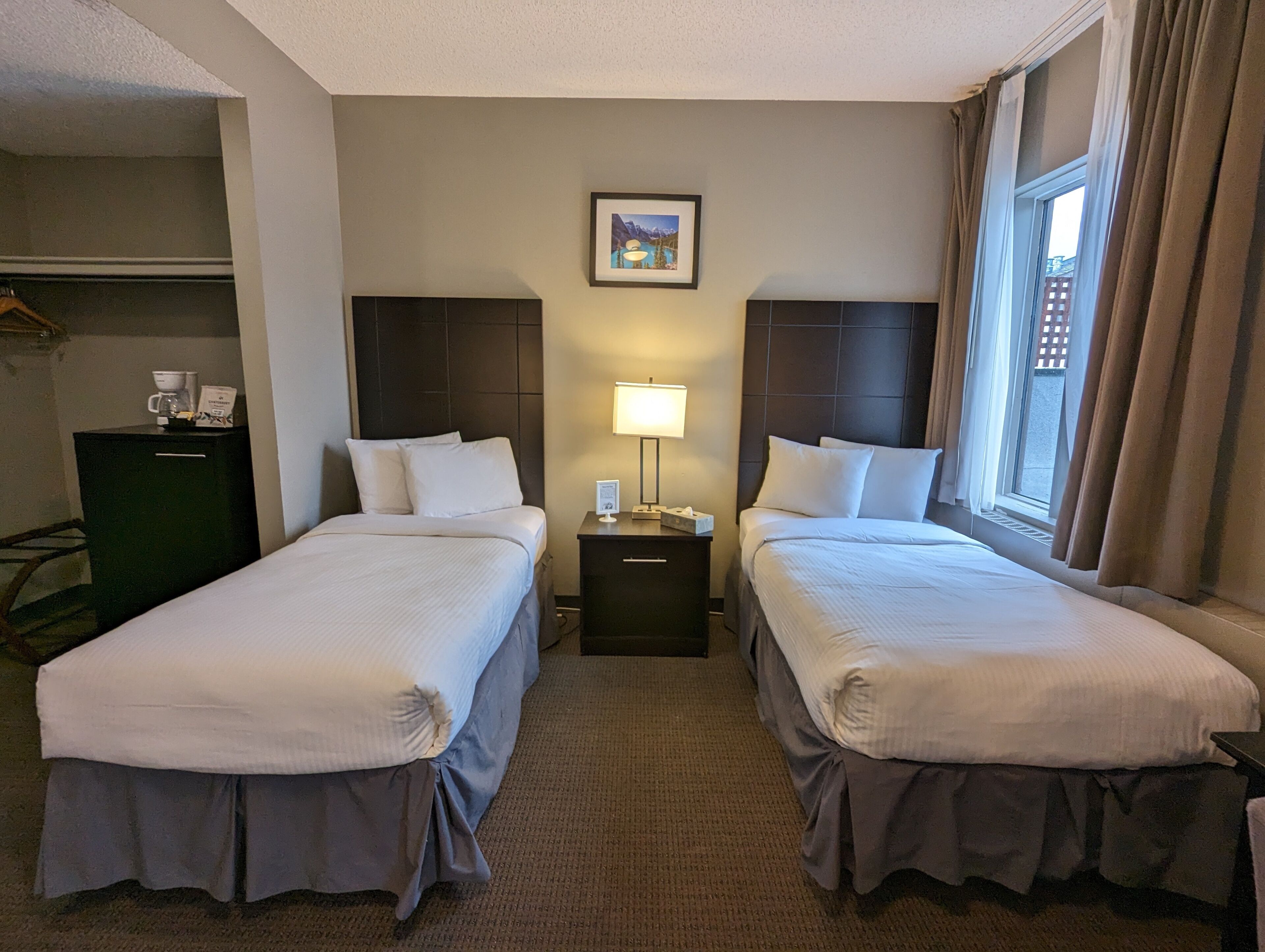 A hotel room with two beds, a nightstand with a lamp, a framed picture on the wall, and a window with curtains.