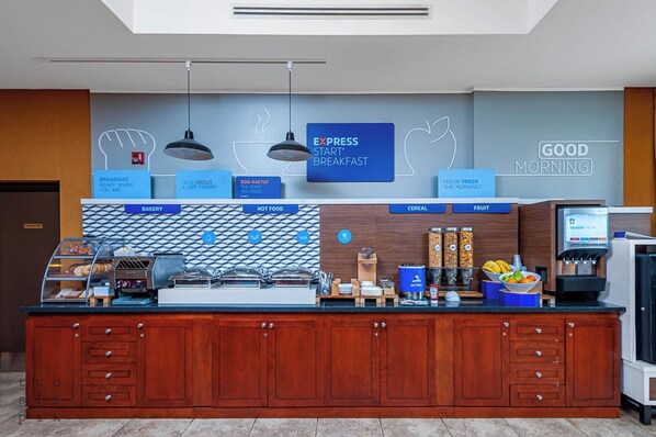 Una barra de desayuno con un letrero que dice "EXPRESS START BREAKFAST" y secciones identificadas como "PANADERÍA", "COMIDA CALIENTE", "CEREALES" y "FRUTA".