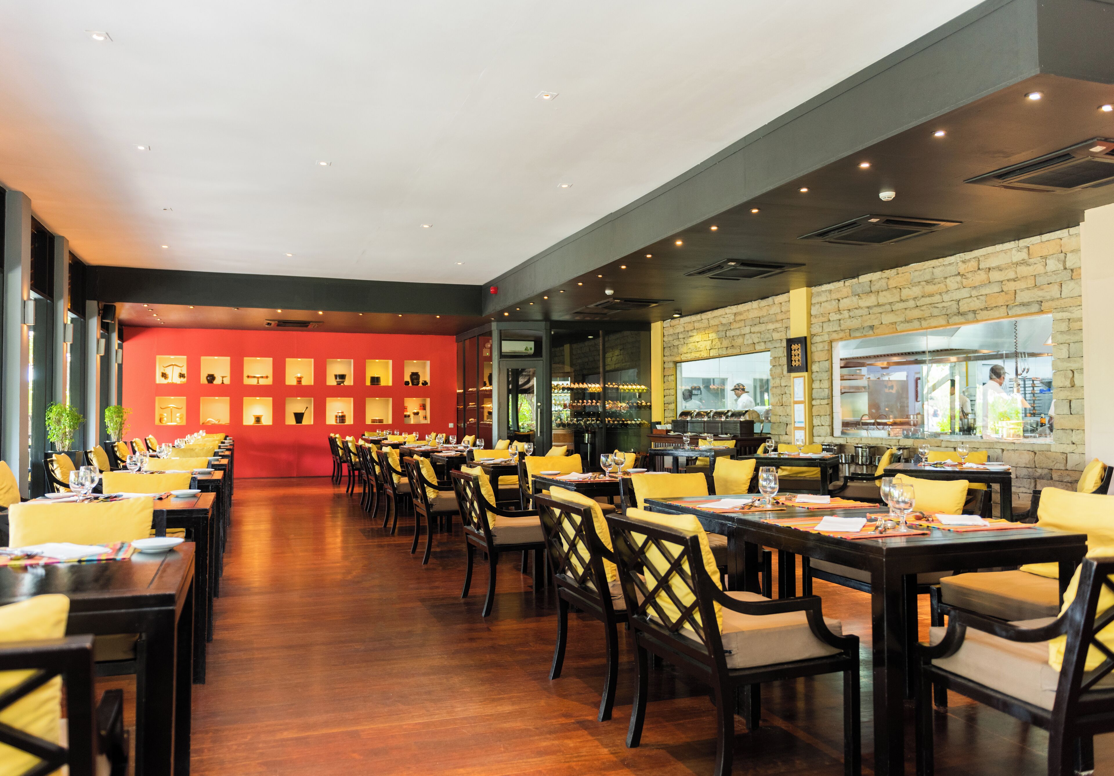Un restaurant avec des tables en bois et des chaises jaunes, un mur rouge orné de tableaux encadrés, et une cuisine visible à travers une cloison vitrée.