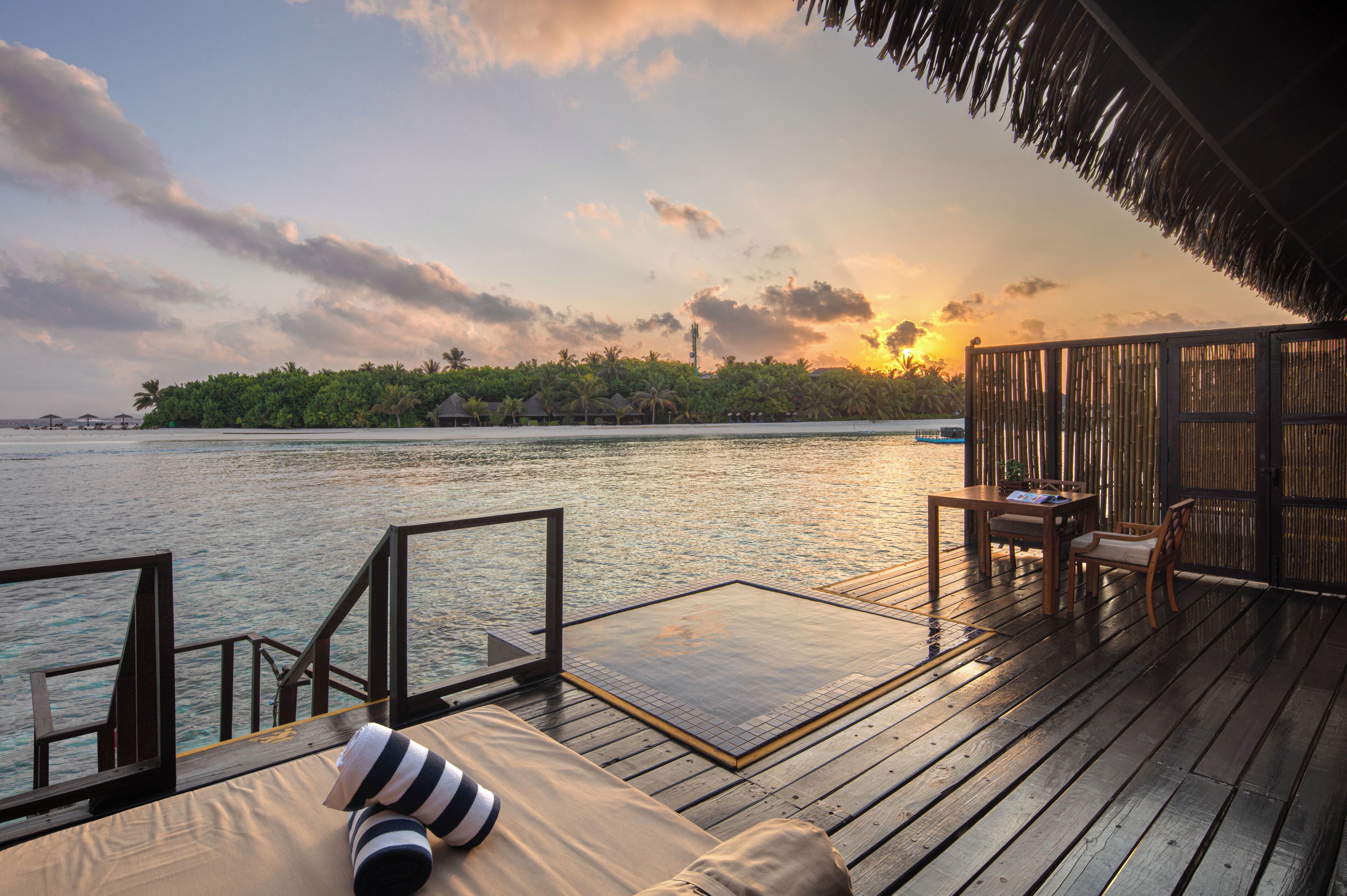 A wooden deck with a sofa and table, overlooking a body of water with a distant island and thatched-roof structures.