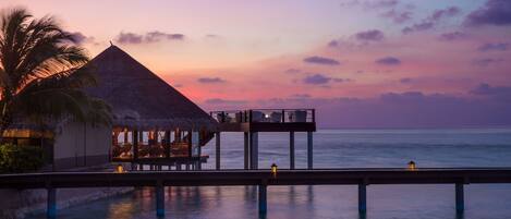 A thatched-roof overwater bungalow with a pool and lounge area, connected to a pier extending into the ocean.