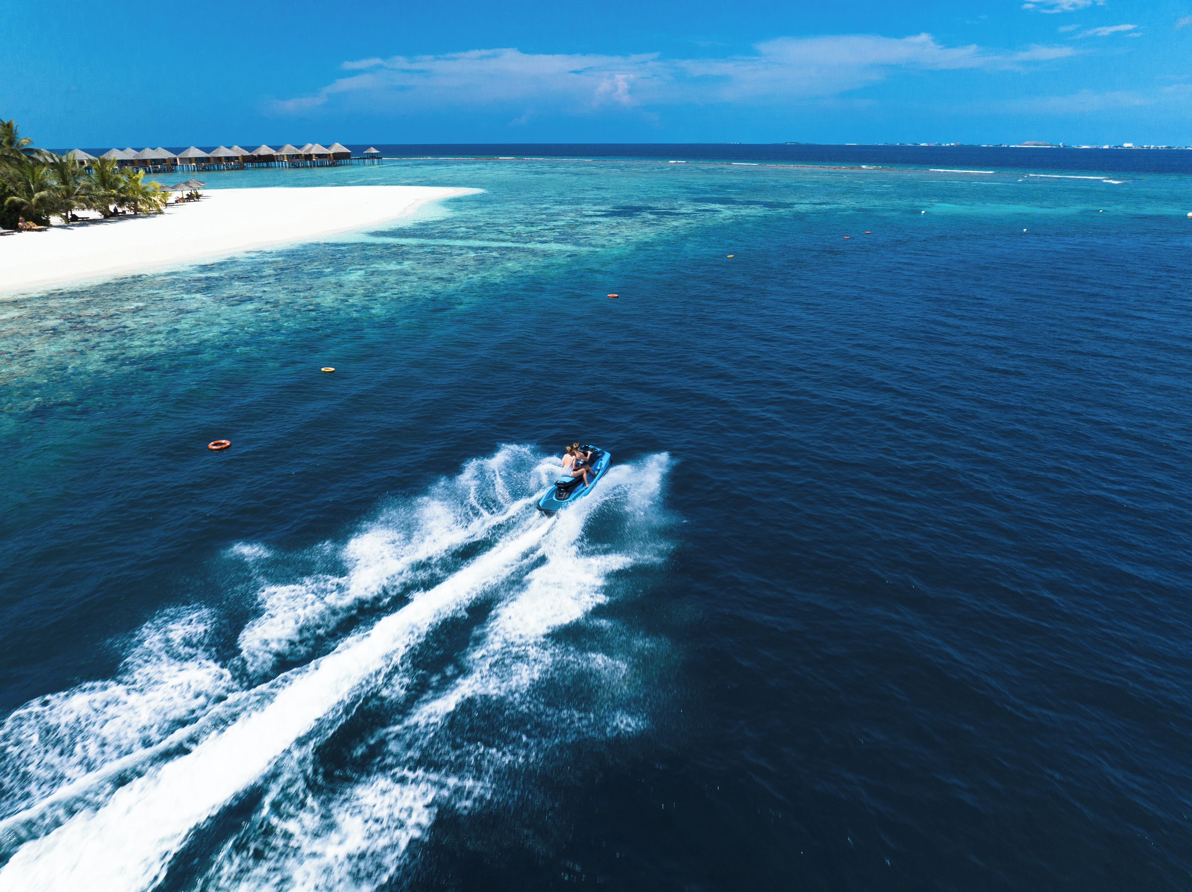 A clear view of a beach with a jet ski in the water, a white sandy shore, and overwater bungalows in the background.