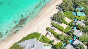An aerial view of a resort with a beach, clear turquoise waters, and several buildings with umbrellas.